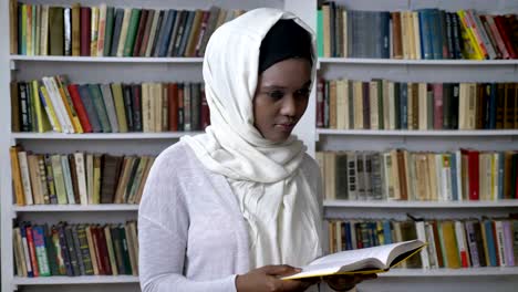 young african muslim girl in hijab is reading book, watching at camera, religioun concept, booksheves on background