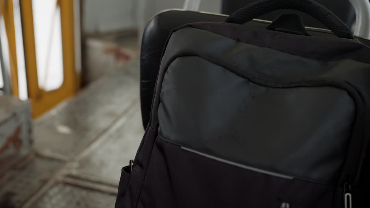 Zaino nero su scale metalliche, primo piano in una stazione urbana che mostra cemento usurato, ringhiera gialla e piattaforma piastrellata, zaino in nylon con tasca con cerniera e manico imbottito, luce mattutina suggestiva, pendolari