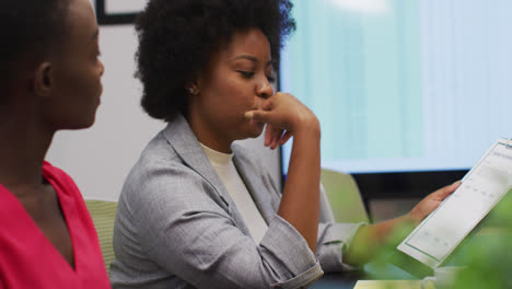 Two-african-american-businesswomen-talking-at-office-meeting,-one-holding-paperwork