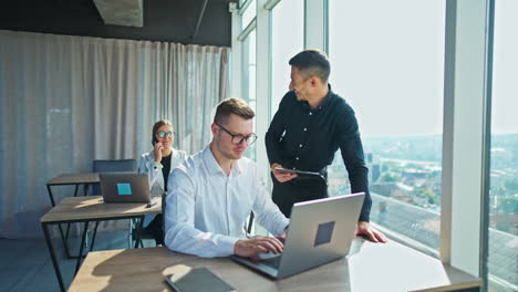 Male colleagues communicate looking at laptop. Man in black shirt turns back to female colleague smiling to her. Sunny office background.