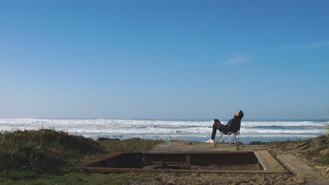 Man-sitting-in-folding-chair-at-the-beach-and-watches-the-waves-in-the-ocean-and-thinking-about-life---relaxing-holiday---freedom---alone-and+d-introverted,-4k-footage-slow-motion