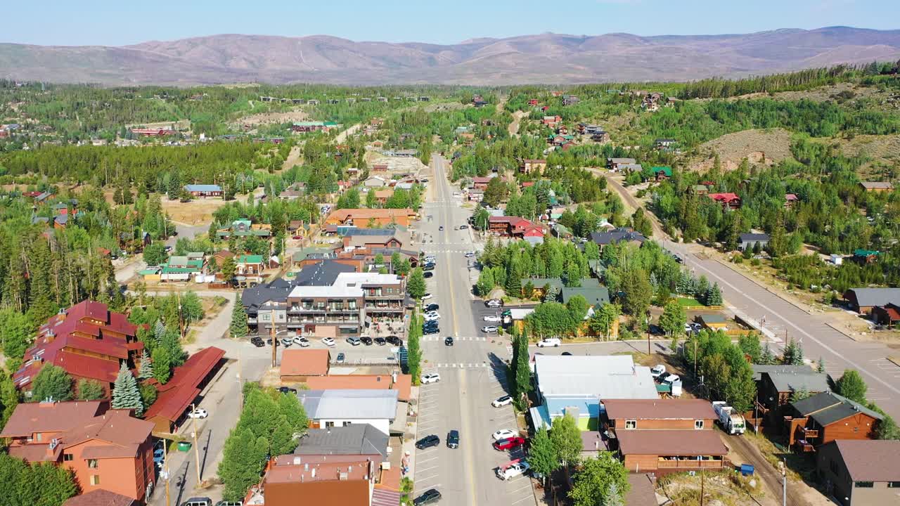 Ripresa con drone sopra una comunità alpina lacustre che rivela una strada centrale, edifici sul lungolago, quartieri boscosi e ampi pendii montani sotto cieli estivi luminosi e sereni