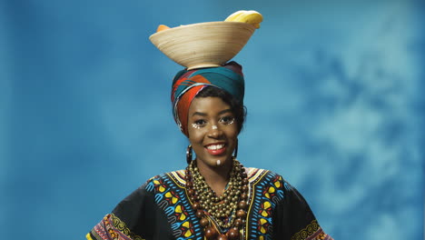 joven afroamericana alegre con turbante y maquillaje sonriendo alegremente a la cámara mientras sostiene un plato con frutas en la cabeza