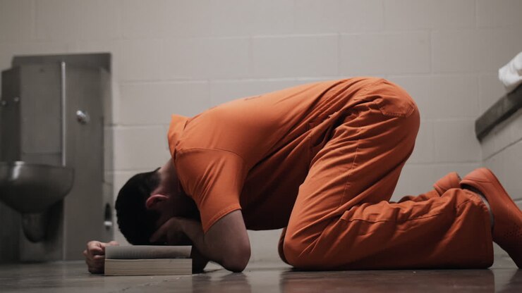Un detenuto triste, un uomo in carcere che indossa una tuta arancione da prigioniero, in ginocchio, piange con una Bibbia, pregando, simbolo di religiosità, fede in Dio, cristianesimo
