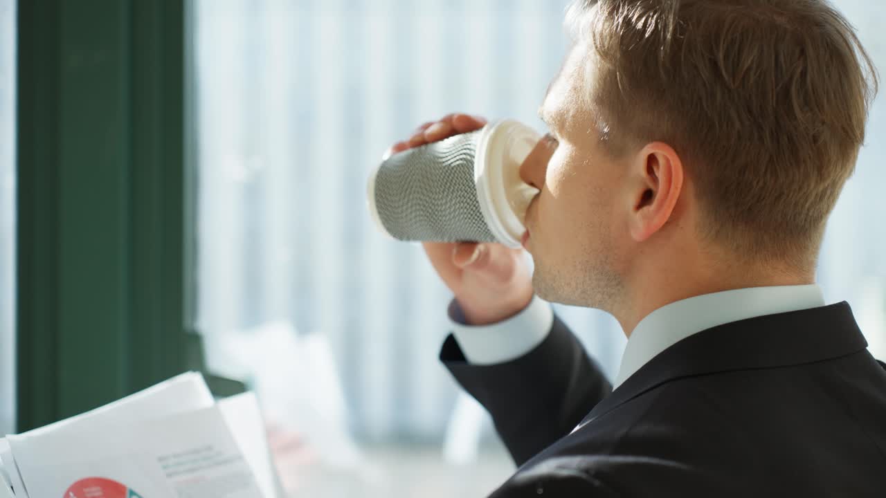 A Caucasian Businessman drinking coffee and reading a report by a window in an office setting daytime