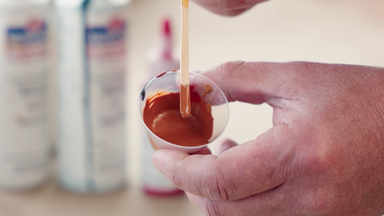 Static view shows craftsman stirring brown paint in cup, testing hue