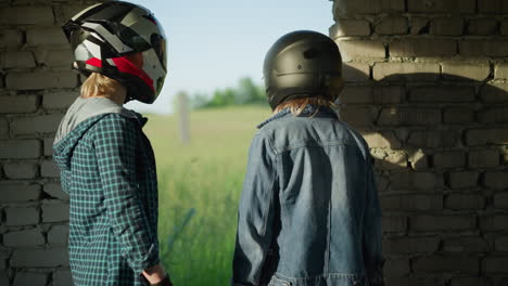 two friends wearing helmets stand inside an abandoned building looking toward a field, one friend faces the field while the other looks sideways at her companion