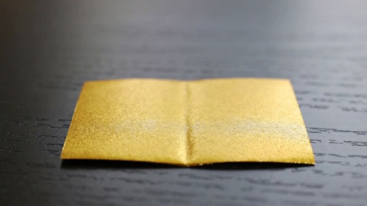 Folding gold foil square guided by central crease, forming standing heart on dark wood table