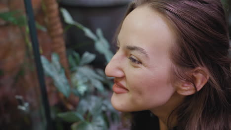 blond woman leans forward with gentle smile and focused expression while observing lush indoor greenery, soft lighting illuminating her face and background filled with textured foliage