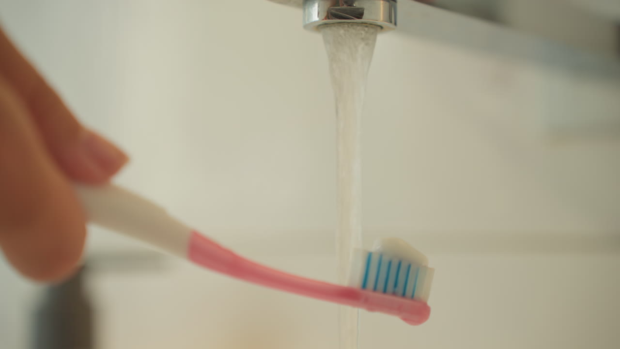 Primer plano de un cepillo de dientes enjuagándose bajo el grifo abierto del lavabo del hotel. Una mano sostiene el mango rosa bajo un chorro claro, con gotas de agua formándose en las cerdas azules. Se aprecian salpicaduras y goteos suaves, creando una atmósfera de despertar matutino para el huésped de viaje.