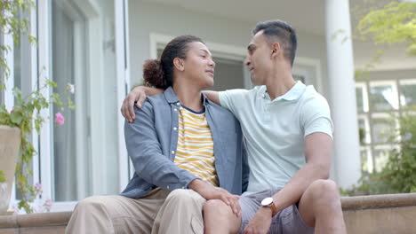 Portrait-of-diverse-gay-male-couple-embracing-and-smiling-sitting-in-sunny-garden,-slow-motion