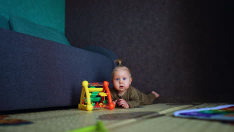 little baby lies on the floor near the sofa. cute blue-eyed kid has a toy in front of him but looks up with interest.