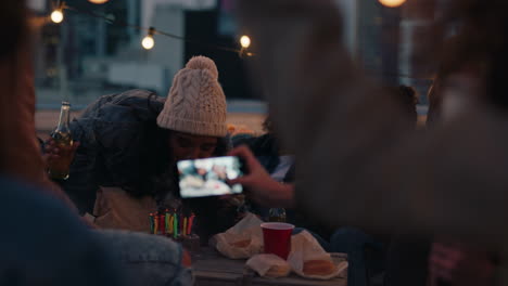 행복한 친구 그룹이 해가 지는 지붕에서 생일 파티를 축하하고 아름다운 젊은 여성이  ⁇ 불을 불어 축하를 즐기고 우정에 토스트를 하고 있습니다.