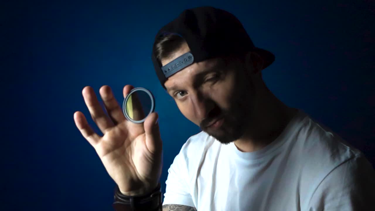Man with white shirt black reversed cap hat and tattoos holds up a CPL polarizer filter and looks through it with confidence and happiness in slow motion while being in front of a blue studio wall