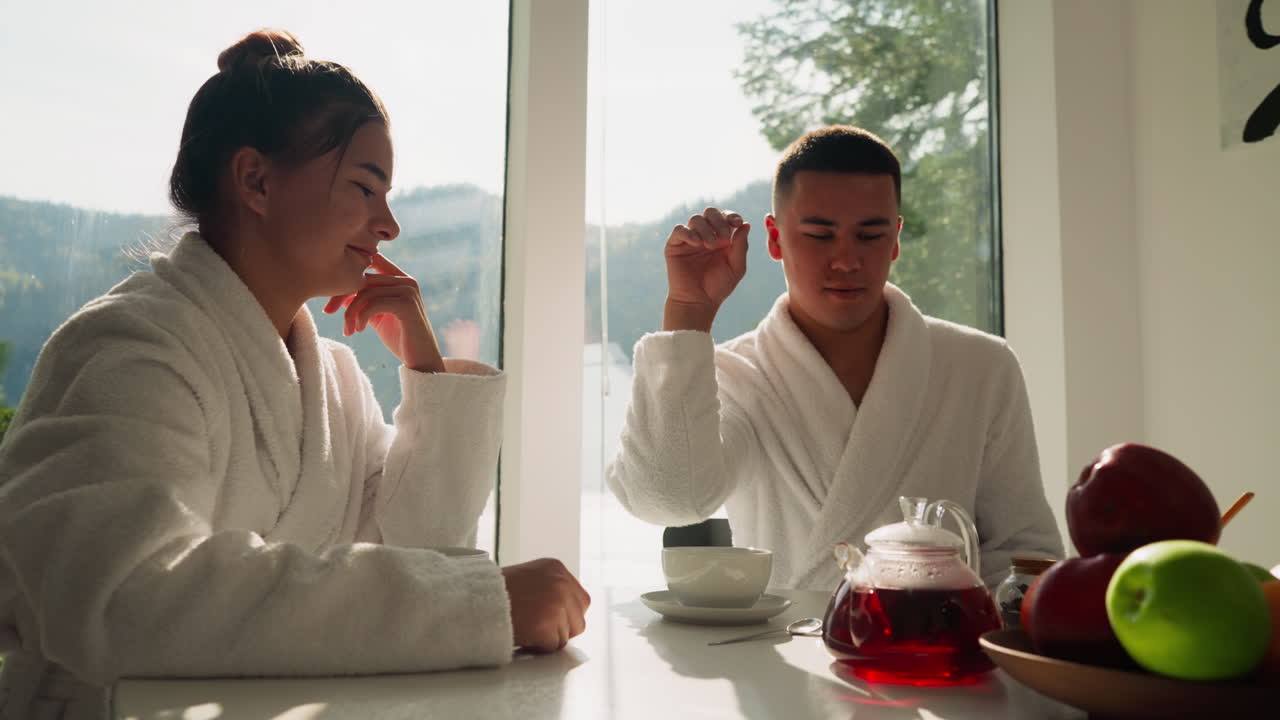 남자가 컵에 건조한 꽃을 던지고 테이블에 앉아있는 여자와 함께 남자친구는 여자친구를 즐겁게하고 아침에 차를 마시며 행복한 커플 생활을합니다.