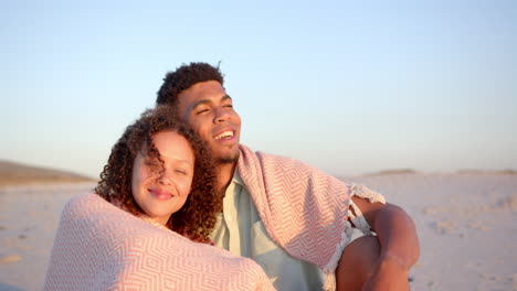 biracial couple wrapped in a blanket, enjoying a beach at sunset