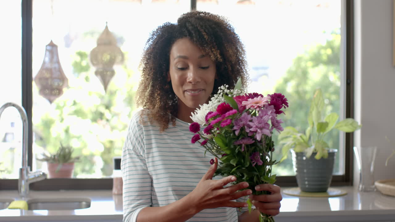 feliz mujer biracial con regalo y flores haciendo una llamada de video en la cocina, en cámara lenta
