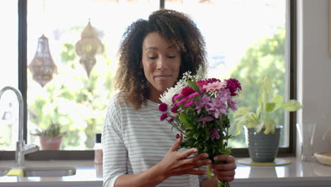 Glückliche-Biracial-Frau-Mit-Geschenk-Und-Blumen-Beim-Videoanruf-In-Der-Küche,-Zeitlupe