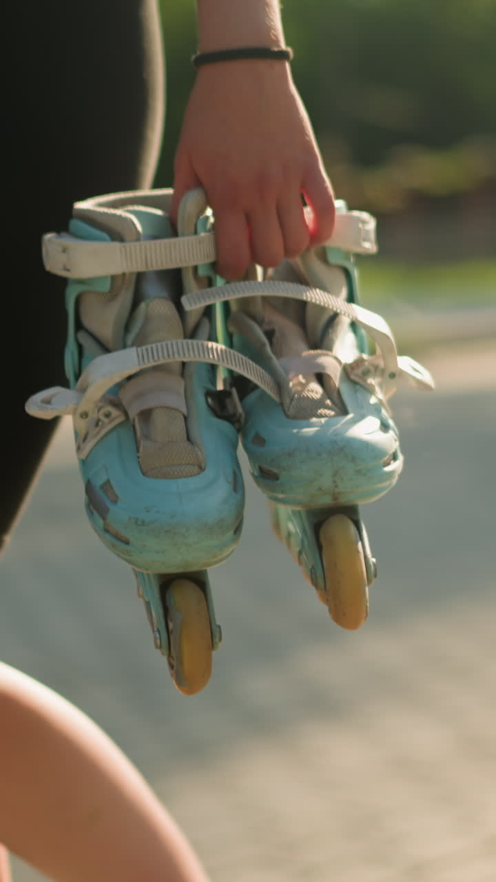 Lady wearing black tights walking outdoors holding cyan roller skates in her right hand on bright sunny day, background features lush greenery and blurred figures of people walking