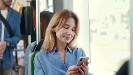 Primer-Plano-De-La-Joven-Mujer-Atractiva-Tocando-Y-Enviando-Mensajes-De-Texto-En-El-Teléfono-Inteligente-Y-Sonriendo-Mientras-Se-Va-A-Casa-En-Tranvía