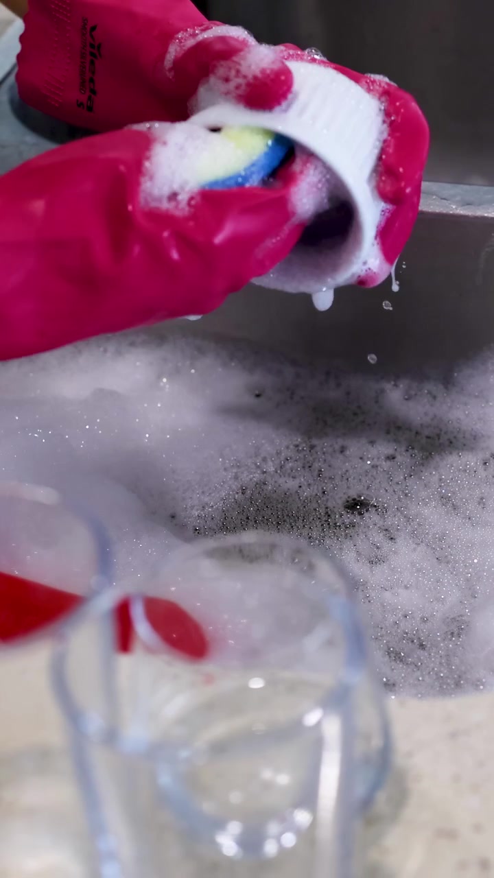 Hands in pink gloves wash a bowl with a sponge in a kitchen sink filled with soapy water