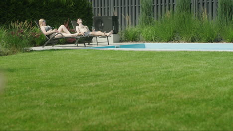 a young couple is relaxing by the pool in the backyard of the house. in the foreground, a well-groomed green lawn