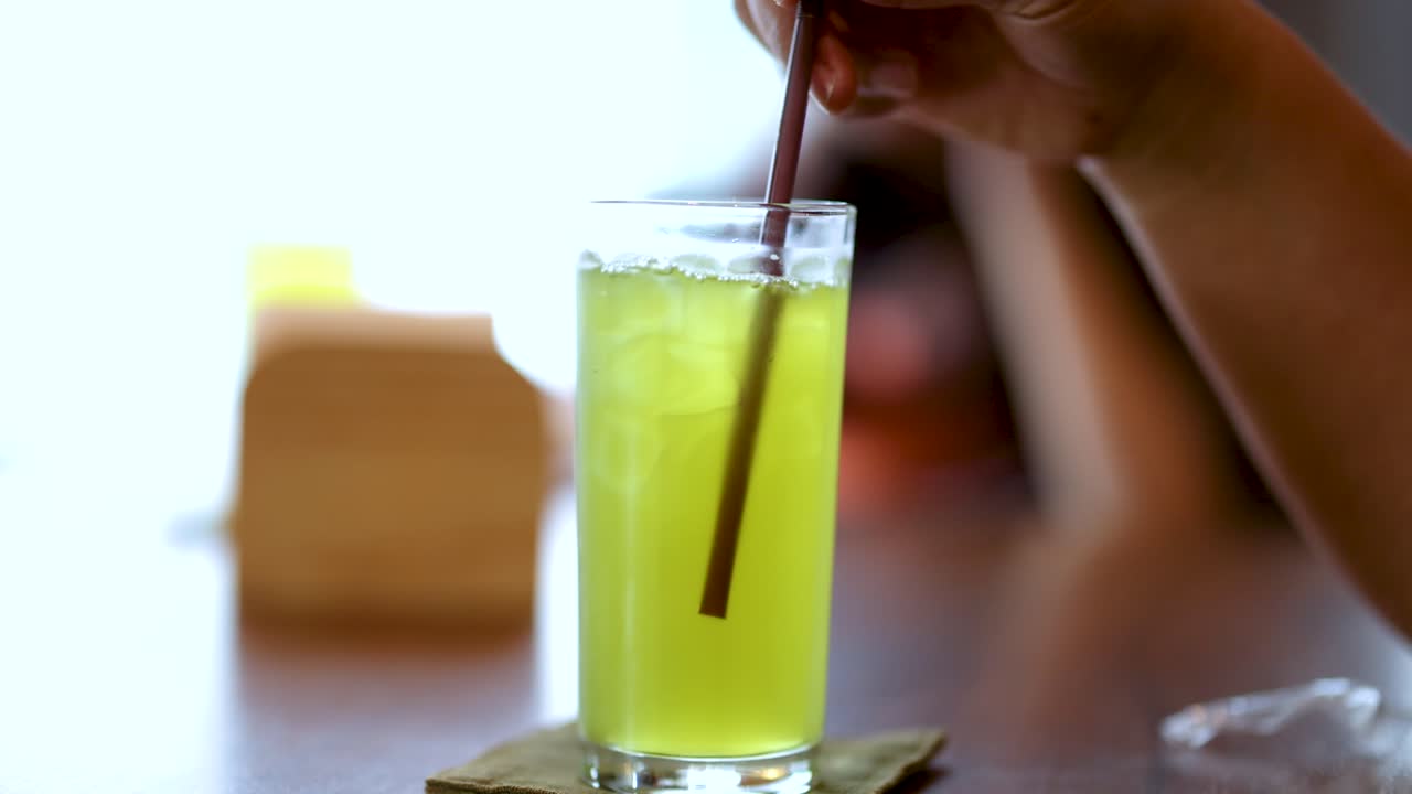Hand stirs iced green tea in glass, natural daylight, shallow focus, relaxed dining environment