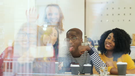 camera focuses on a multiethnic group of friends through the window making a selfie and doing funny gestures while they are sitting at a table in a cafe