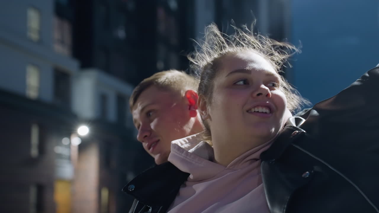 Close up of cheerful siblings dancing under street light with wind swaying lady hair, joyfully smiling and enjoying moment outdoors with tall residential building glowing in background
