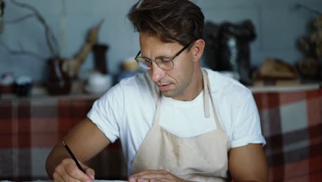 Ein-Glücklicher-Junger-Töpfer-Mit-Brille-Und-Weißem-T-Shirt-Schreibt-Seine-Pläne-Mit-Einem-Bleistift-Auf-Papier.-Traditionelles-Geschäftskonzept