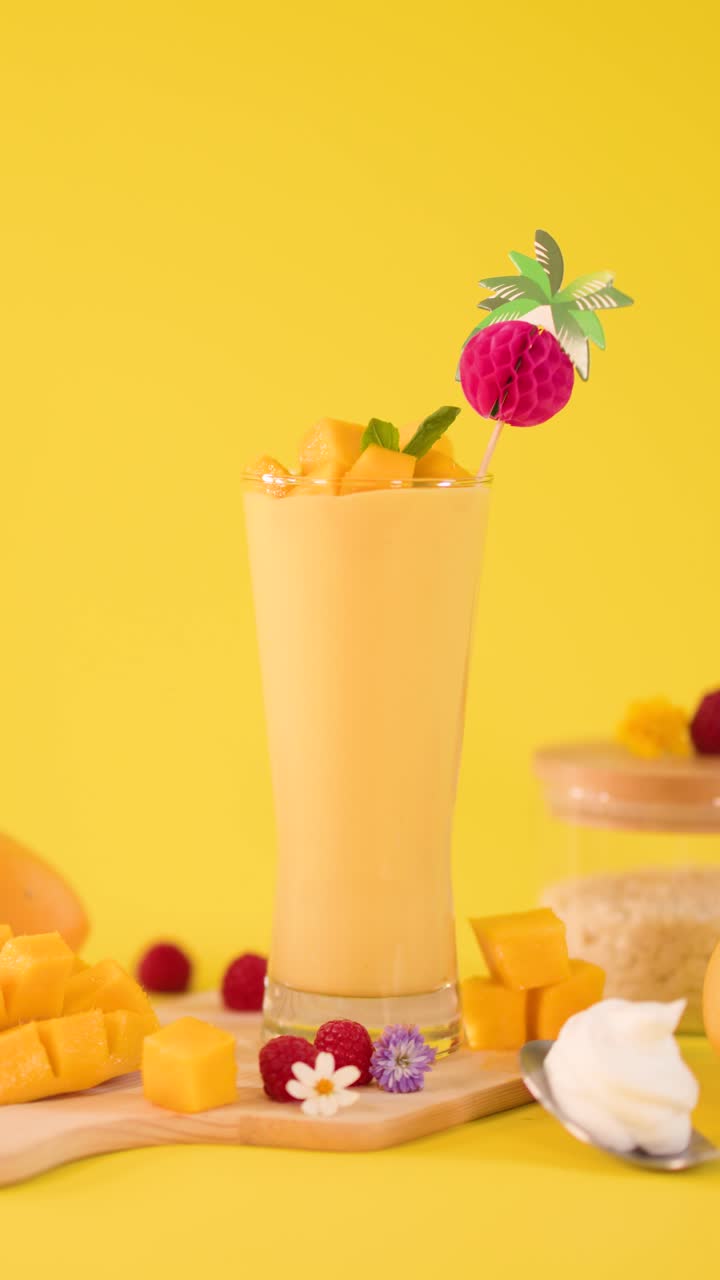 Primo piano statico di frullato di mango su sfondo giallo mentre una mano aggiunge una guarnizione tropicale giocosa