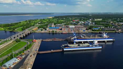 puerto industrial con buques atracados, almacenes e infraestructura circundante cerca de una vía fluvial y zonas verdes exuberantes en letonia