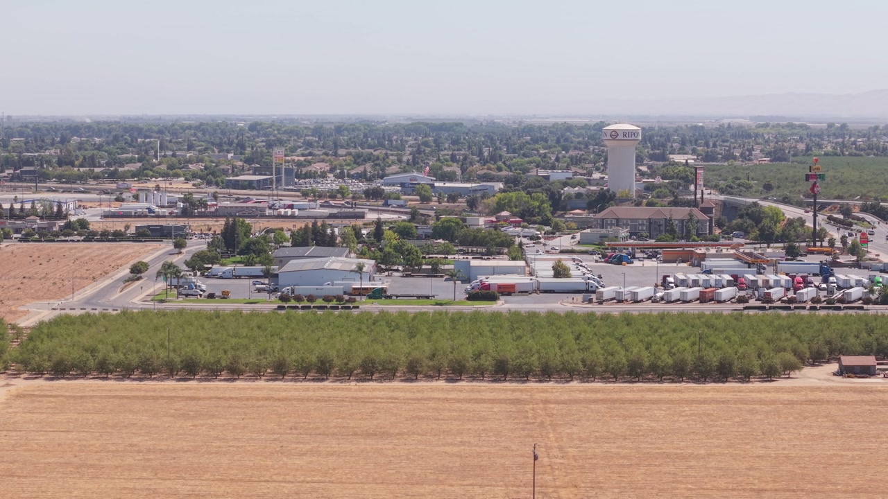 Side aerial shot along field with rows of trees, parking full of trucks, water tower, and busy road leading to Ripon California city.