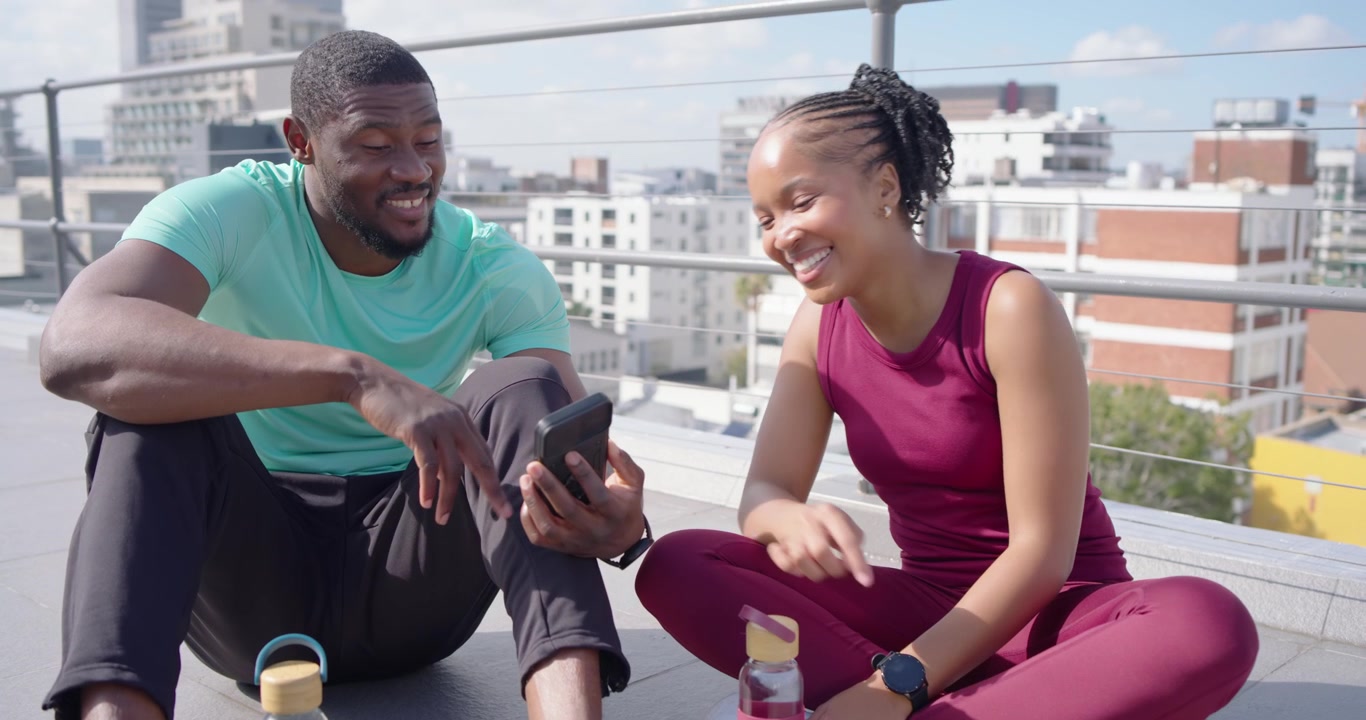 African couple in sportswear sharing laugh on rooftop, man tapping phone, woman sipping pink bottle