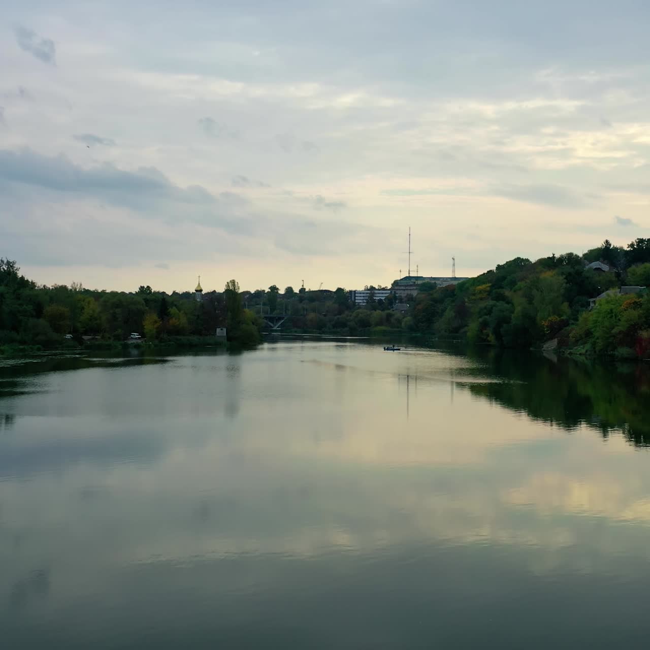 Calm water in the evening. River among green trees from both sides. Panoramic view of a river in the town. Motion camera back. Aerial view.