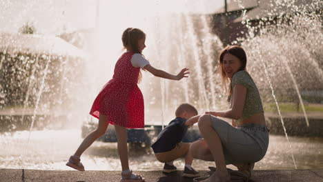 girl splashes water in mother squatting with son by fountain