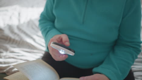 senior woman in teal sweater sitting on bed, holding a magnifying glass to read a hardcover book. soft light shines on the scene, creating a tranquil and focused atmosphere