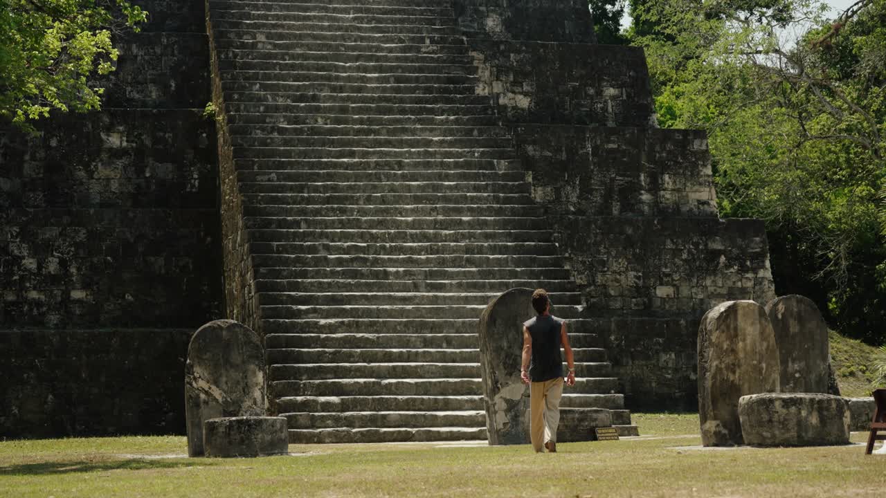 A barefoot man stands before a towering ancient Mayan pyramid in Tikal, surrounded by carved stone stelae and dense jungle.