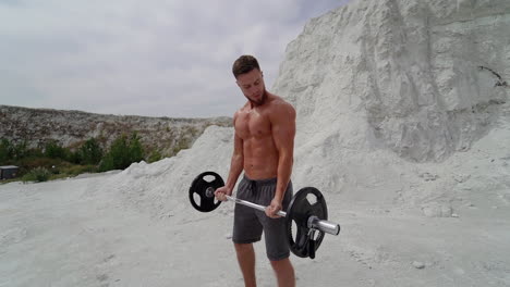Muscular shirtless man exercising hard in the mountains. Bodybuilder with barbell on white mountain background in summer outdoors.