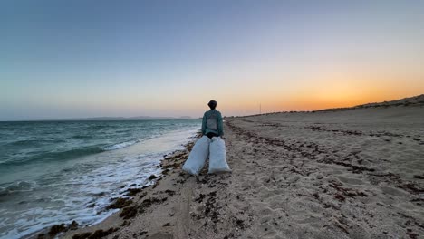 Eine-Frau,-Die-Im-Sommer-Am-Wunderschönen-Sandstrand-Am-Meer-Entlang-Läuft-Und-Einen-Müllsack-Bei-Sonnenuntergang-Trägt,-Während-Sie-Die-Meeresverschmutzung-In-Einer-Malerischen-Panoramalandschaft-Der-Iranischen-Natur-Säubert