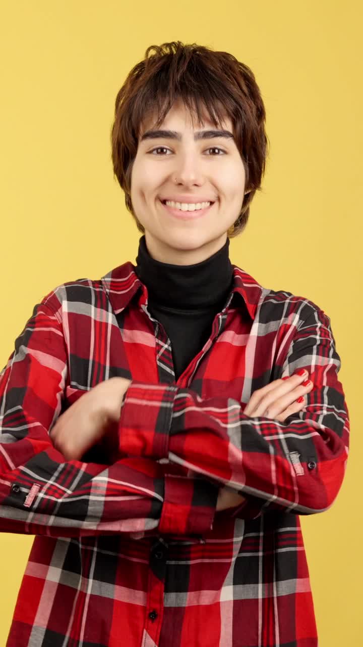 Smiling young woman with short hair wearing a red plaid shirt and a black turtleneck against a yellow background