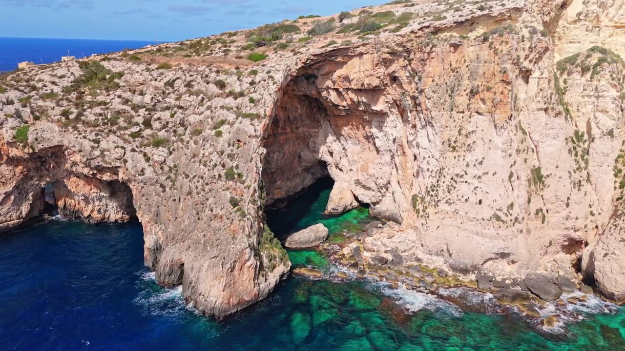 Maltas Blaue Grotte, ein majestätischer natürlicher Kalksteinbogen und Höhlenkomplex am Meer an der Südküste. Weitläufige Luftaufnahmen zeigen tief azurblaues Wasser, das mit schroffen, sonnenverwöhnten Klippen kontrastiert.