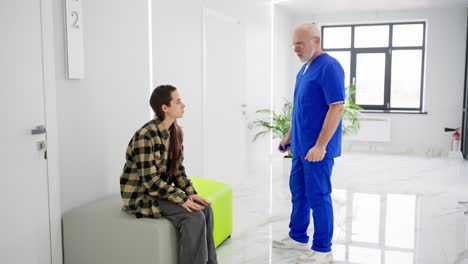 un médico de edad avanzada confiado con barba y uniforme azul se comunica con una joven morena con una camisa a cuadros sobre su problema y diagnóstico en una clínica moderna