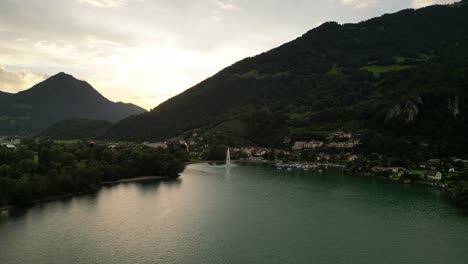 vista de pájaro de grandes montañas escondiendo una pequeña ciudad bajo las colinas junto al agua con una gran fuente salpicando agua en lo alto y un muelle lleno de barcos listos para la aventura lugar relajante para salir