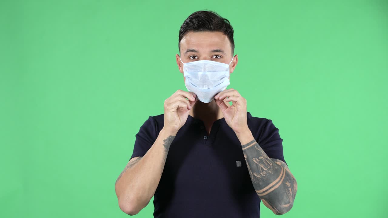 Portrait of a handsome young guy looking at camera and puts on medical mask. Health Protection Corona Virus Concept. Green screen
