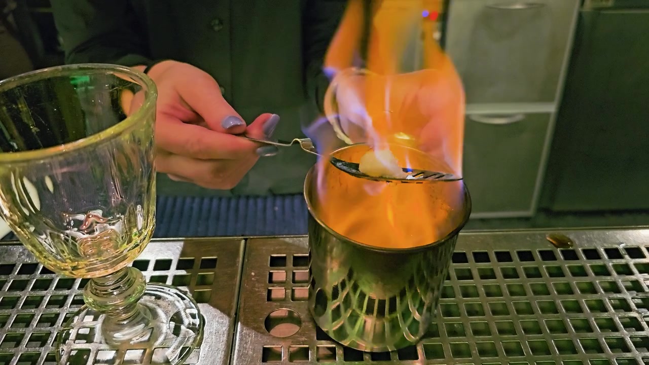 Primo piano di un classico servizio di assenzio in un bar, che mostra una fiamma blu, una zolletta di zucchero su un cucchiaino, una tazza di metallo, un bicchiere a calice, le mani del barista e l'allestimento del bancone in acciaio inossidabile riflettente.