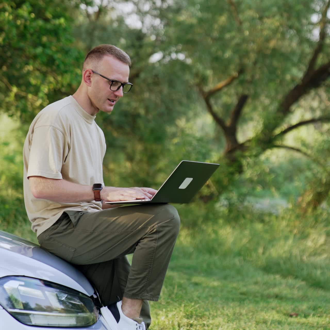 Caucasian male sitting on the car front holding a laptop. Freelancing man types on computer and smiles