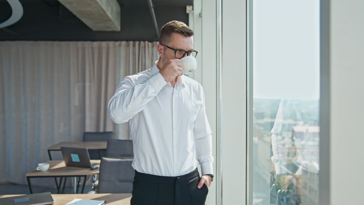 Businessman drinking coffee standing in the spacious office. Man enjoys coffee break looking at the city from a big panoramic window.