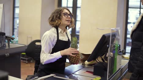 Young-Curly-Woman-Cashier-Is-Kindly-Talking-To-The-Customer-From-The-Register-Counter,-Then-Keep-Scanning-Products,-Packing-It-Into-Bag