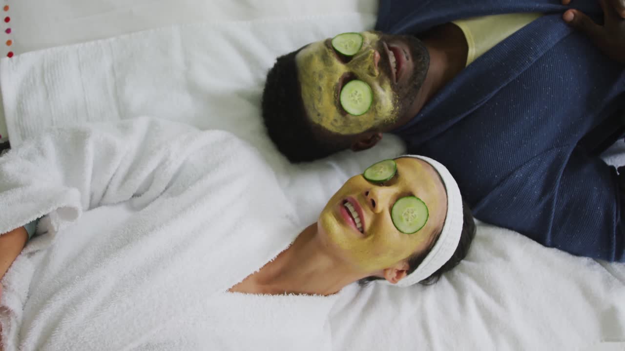 Happy diverse couple lying with masks and pieces of cucumber on eyes in bathroom